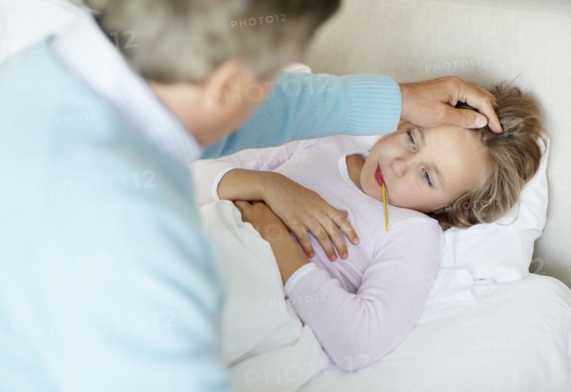 Grandfather tending granddaughter (10-11) lying in bed with thermometer in mouth. Photo : Momentimages