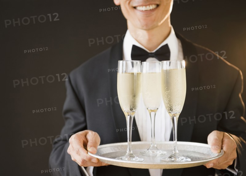 Waiter wearing bow tie and holding tray with champagne flutes, studio shot. Photo : Jamie Grill