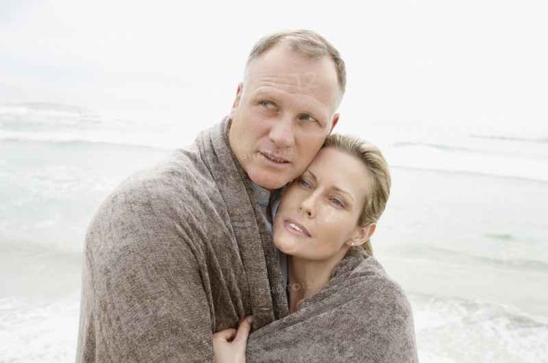 Embraced couple covered by blanket looking away on beach. Photo : Momentimages