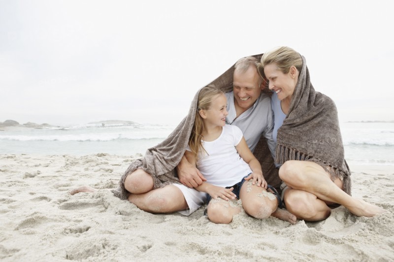 Smiling parents with daughter (10-11) on coastline covered by blanket. Photo : Momentimages