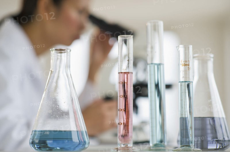 USA, New Jersey, Jersey City, Female scientist using microscope behind test tubes and flasks.