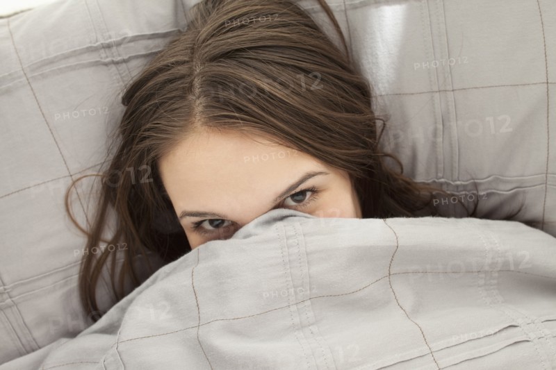 Portrait of young woman lying in bed hiding under bedding. Photo : Mike Kemp