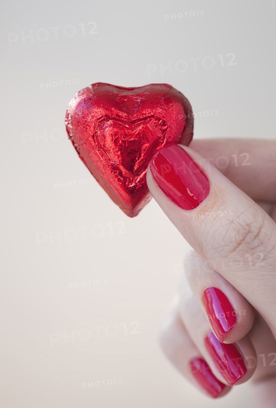 Close up of woman's fingers with red nail polish holding chocolate in shape of heart in red wrapping.
