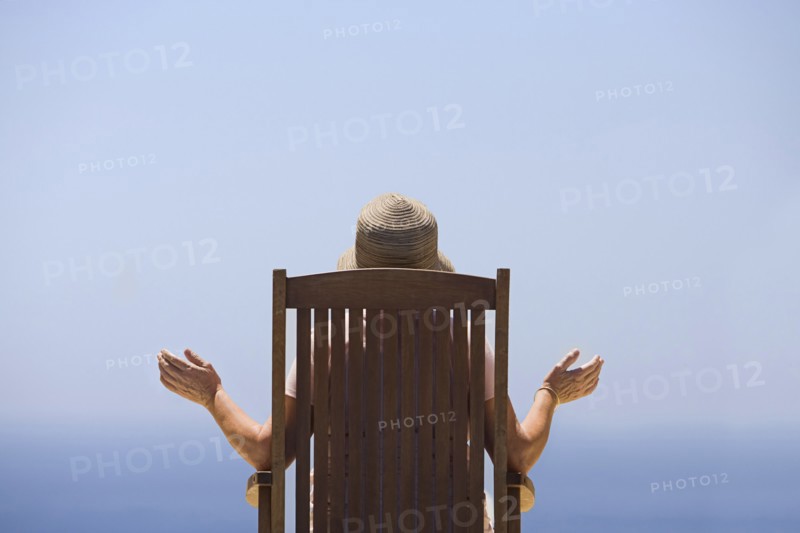 Rear view of woman sitting on wooden chair, facing sea. Photo : Justin Paget
