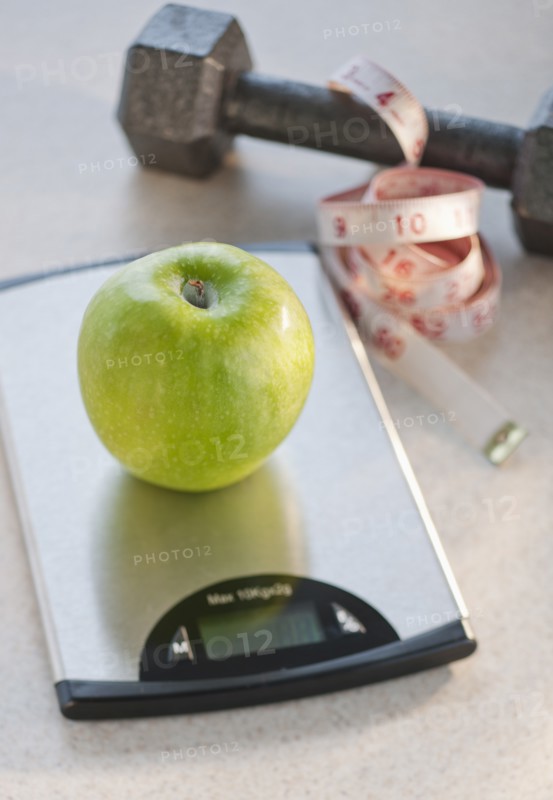 Green apple on weight scale, tape measure and exercise weight in background.