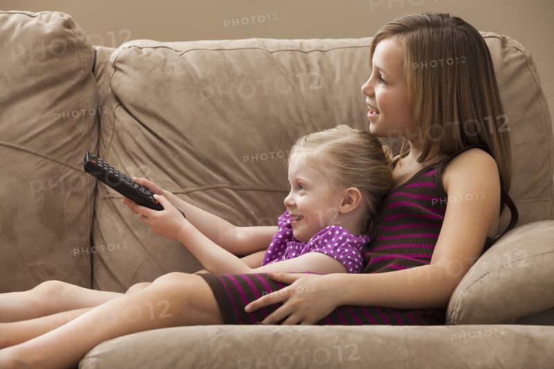 Girl (10-11) with sister (2-3) sitting on sofa, watching TV. Photo : Mike Kemp
