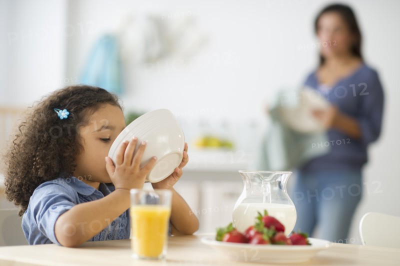 Girl (6-7) eating breakfast with defocused mother in background.