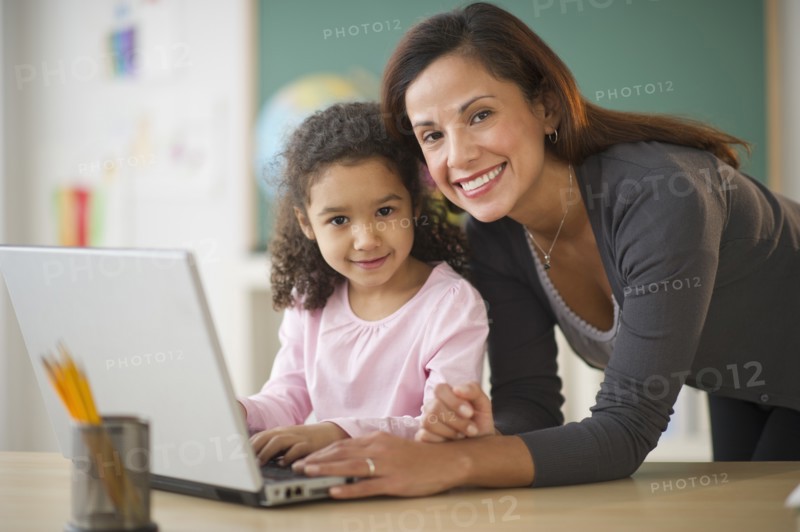 Girl (6-7) with female teacher using laptop in classroom.