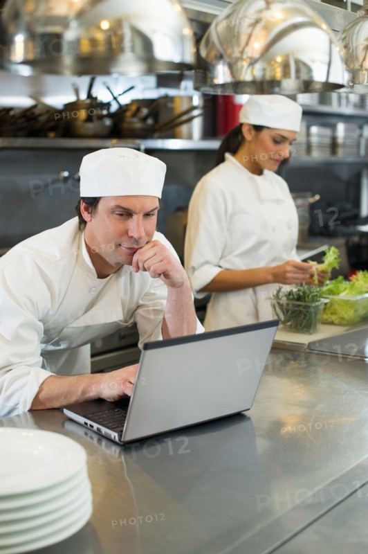 Chef using laptop and cook preparing food in commercial kitchen.