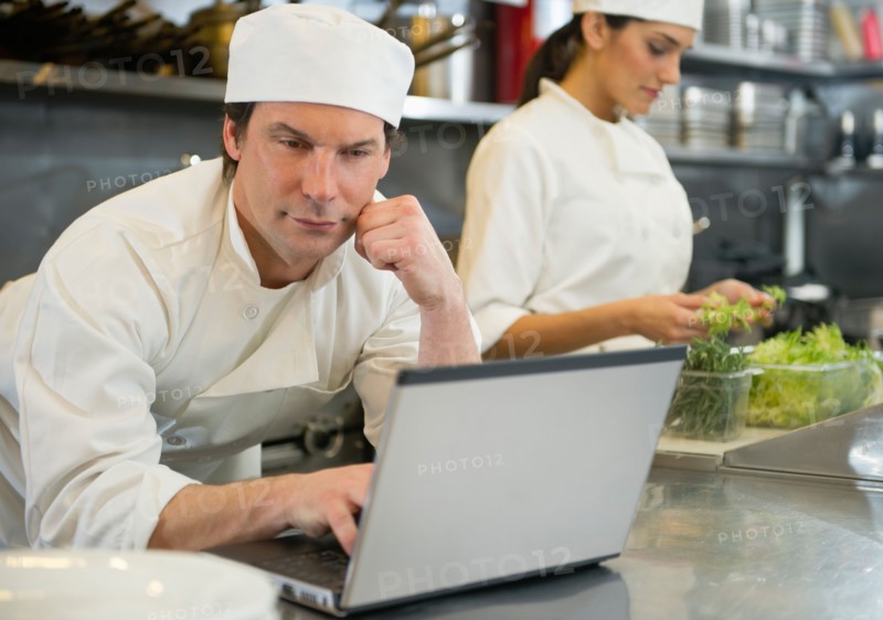 Chef using laptop and cook preparing food in commercial kitchen.