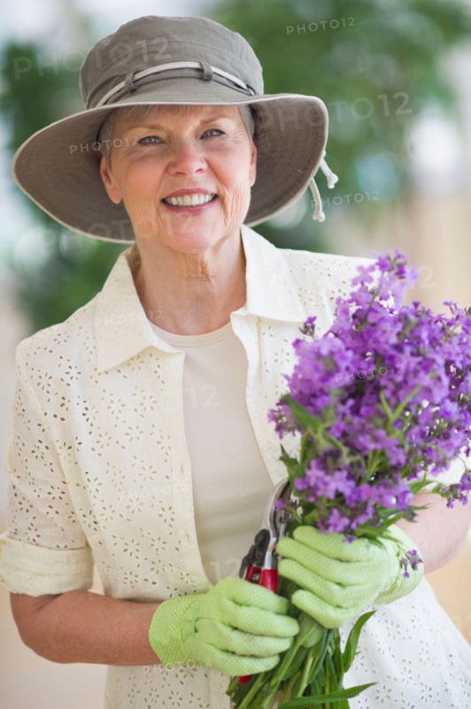 Portrait of smiling senior woman wearing gardening gloves, holding bunch of purple flowers.