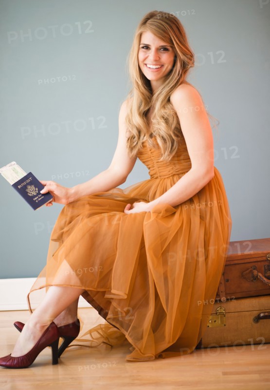Woman in dress sitting on luggage and holding passport. Photo: Jamie Grill