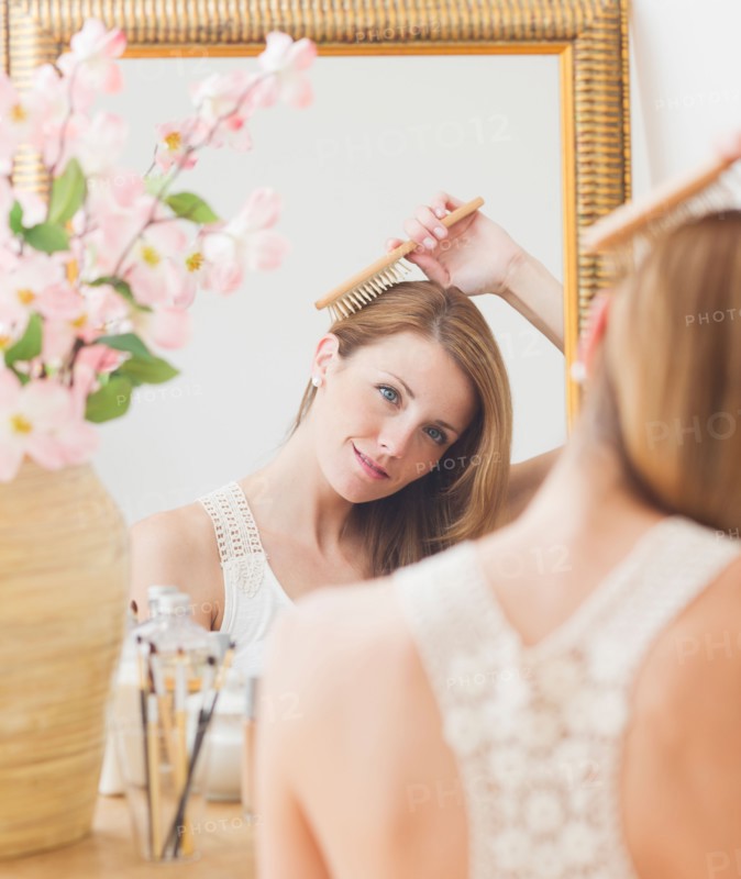 Young woman brushing hair in front of mirror. Photo : Daniel Grill