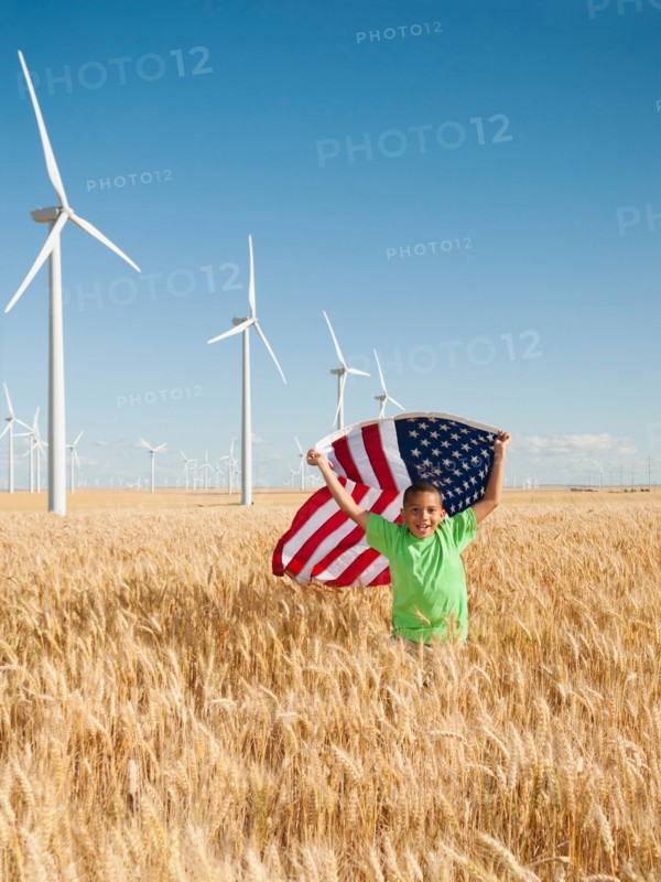 USA, Oregon, Wasco, Boy (8-9) flying american flag in wheat field with wind turbines in background.