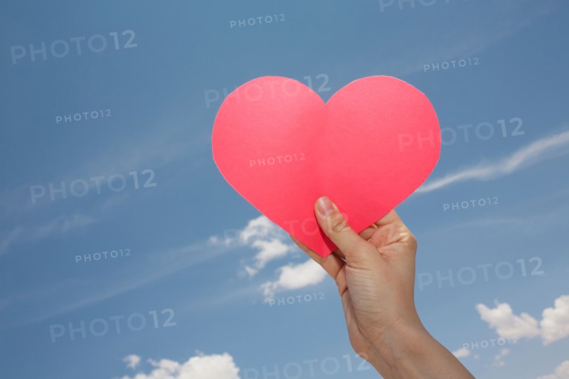 Woman's hand holding pink heart shape against sky. Photo: Winslow Productions