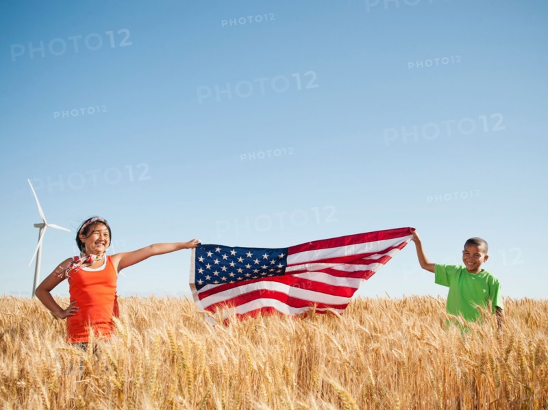 USA, Oregon, Wasco, Girl (10-110 and boy (8-9) holding american flag in wheat field, wind turbine in background.