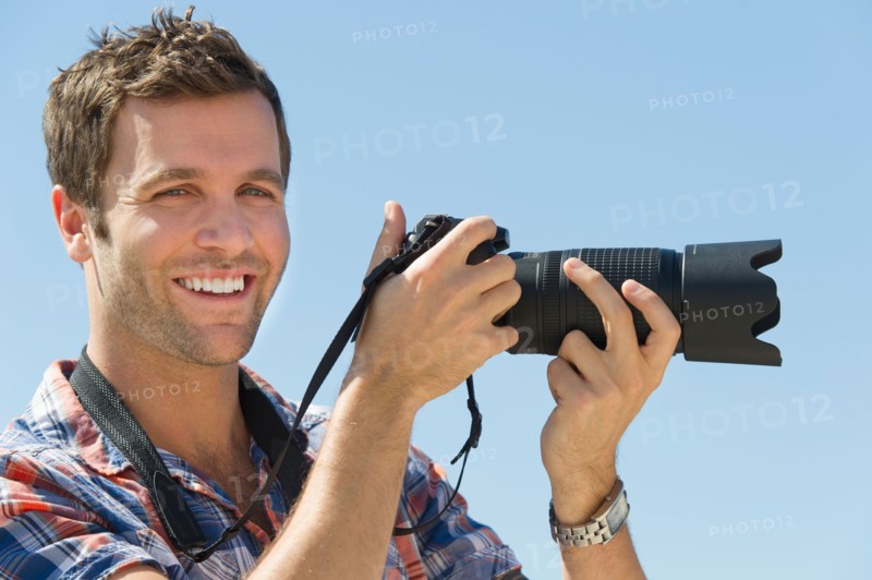 Portrait of man holding camera against blue sky.
