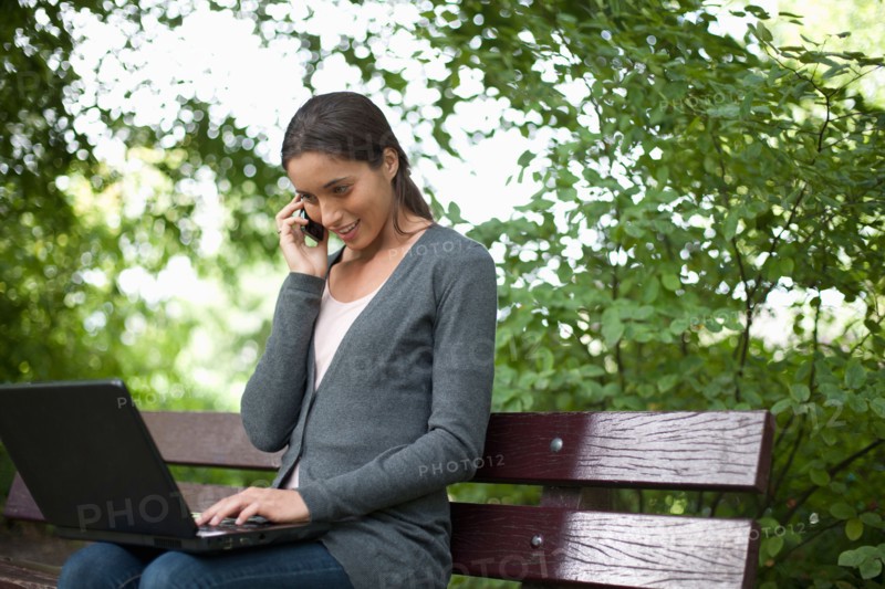 Young woman sitting on park bench using laptop and cell phone. Photo : Jan Scherders