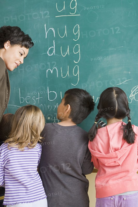 Children (6-7) writing on blackboard with teacher looking at them. Photo: Rob Lewine