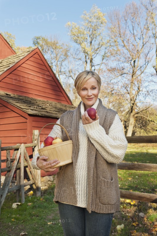 USA, New Jersey, Portrait of smiling woman holding basket with apples in front of cottage house in Autumn. Photo : Tetra Images