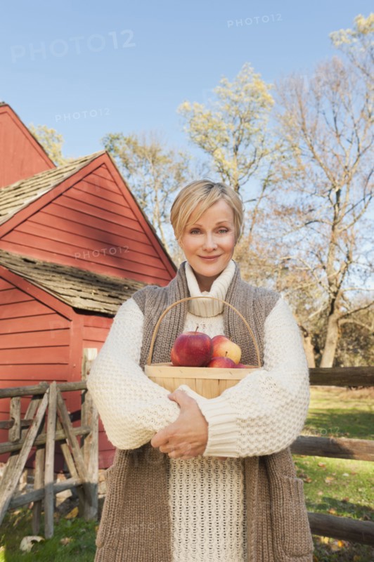 USA, New Jersey, Portrait of smiling woman holding basket with apples in front of cottage house in Autumn. Photo : Tetra Images