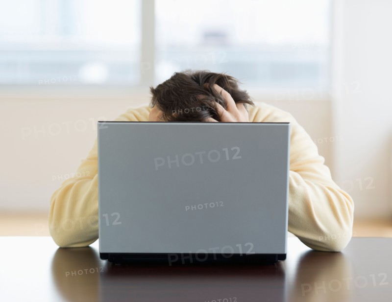 Stressed man sitting at desk behind laptop with head in hands.