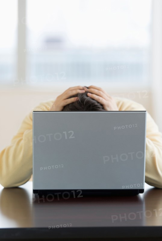 Stressed man sitting at desk behind laptop with head in hands.