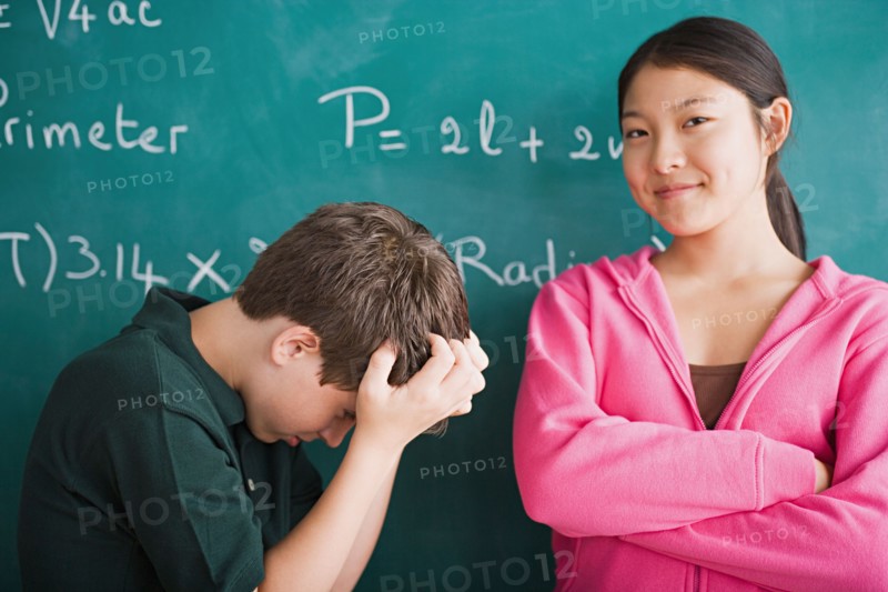 Happy girl and frustrated boy standing in front of blackboard with mathematical formula. Photo : Rob Lewine