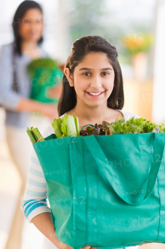 Daughter (12-13) holding bag of fresh vegetables, mother in background.
