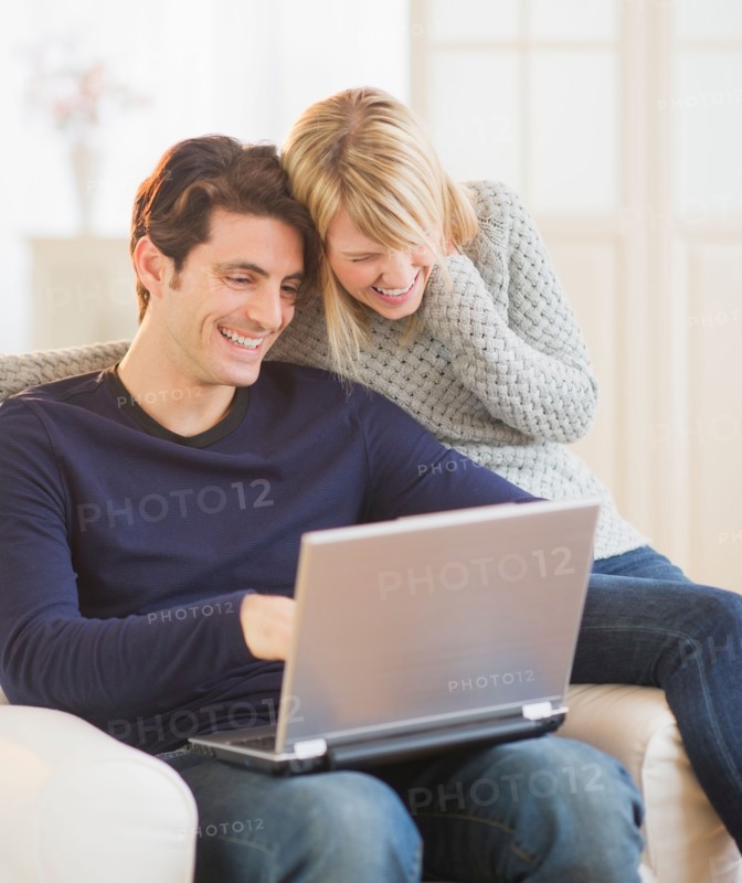 Happy couple sitting together on armchair and using laptop . Photo : Daniel Grill