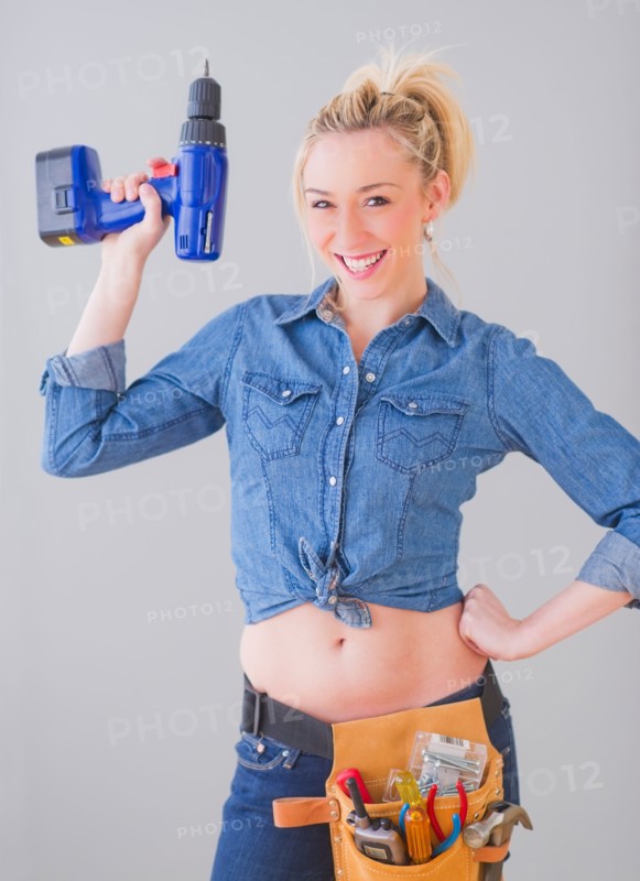 Portrait of young woman wearing tool belt and holding drill, studio shot. Photo : Daniel Grill