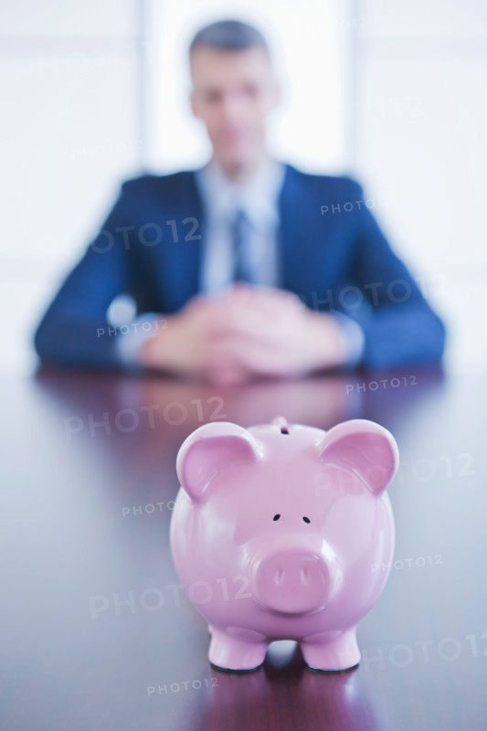 Piggybank on desk, businessman sitting on background. Photo : Daniel Grill