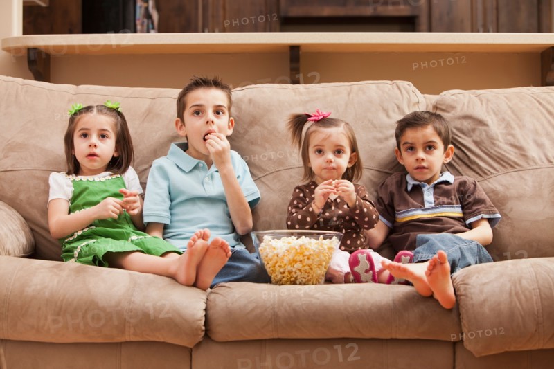 Front view of children (18-23 months, 4-5, 6-7, 8-9) sitting on couch watching tv and eating popcorn. Photo : Mike Kemp