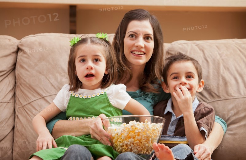 Front view of mother with children (4-5, 6-7) sitting on couch watching tv and eating popcorn. Photo : Mike Kemp