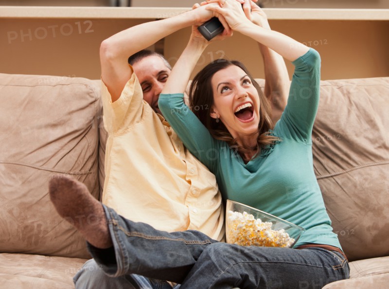 Front view of mid adult couple sitting on couch and fighting for remote control. Photo : Mike Kemp