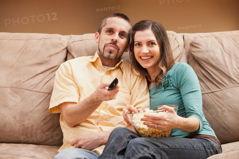 Front view of mid adult couple sitting on couch with remote control and popcorn. Photo : Mike Kemp