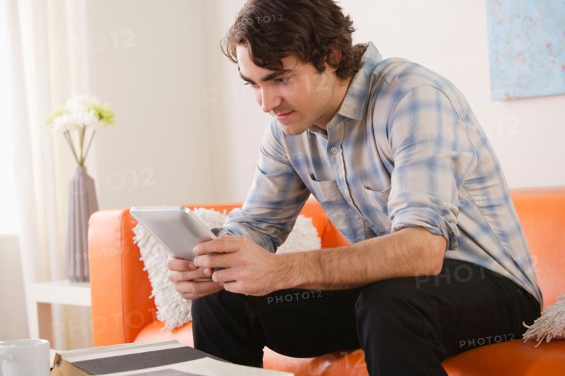Young man sitting on sofa and using digital tablet. Photo : Rob Lewine