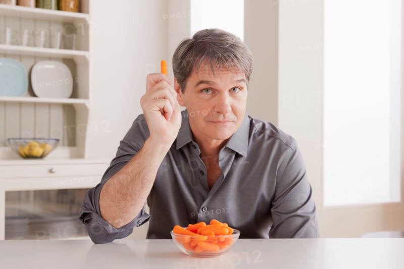 Portrait of mature man with baby carrot on table. Photo : Rob Lewine