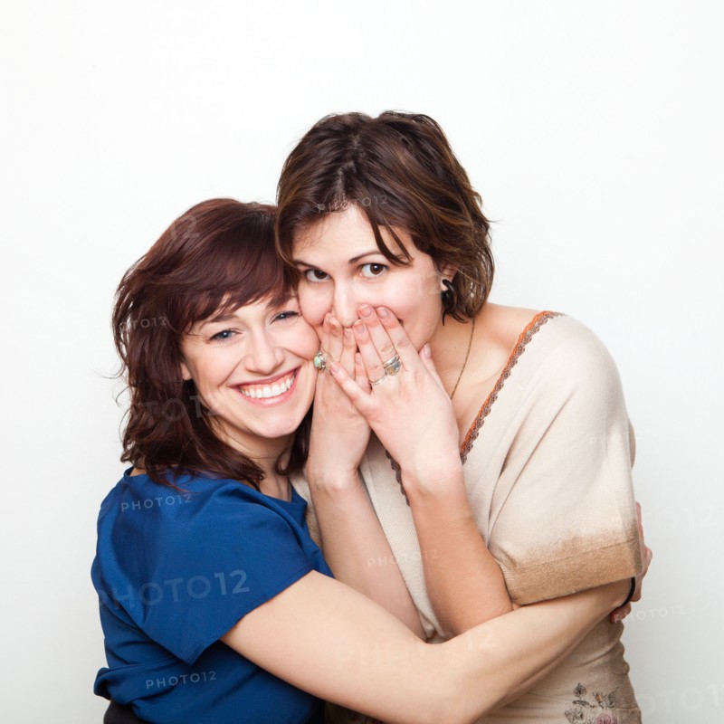 Studio Shot portrait of young women embracing, covering mouth with hand. Photo : Jessica Peterson