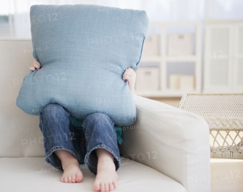 Girl (2-3) sitting on sofa hiding behind pillow. 
Photo : Jamie Grill