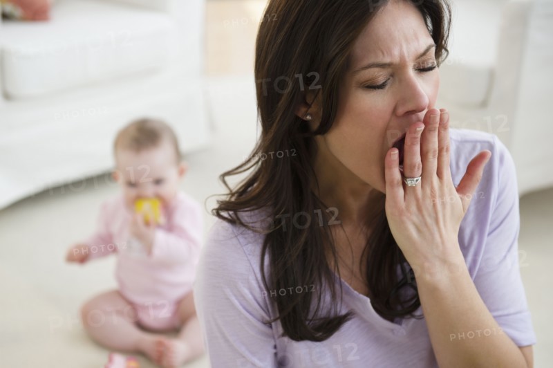 Tired woman yawning, baby daughter (6-11 months) in background. 
Photo: Jamie Grill