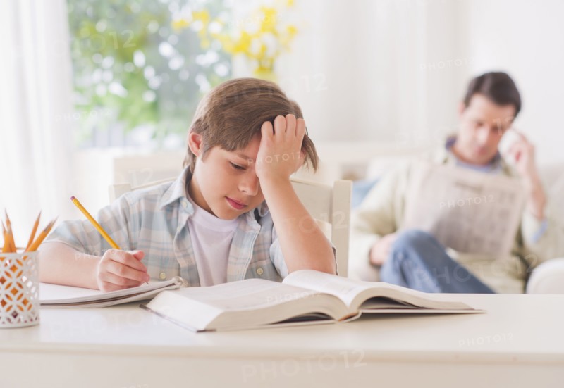 Boy (10-11 years) doing homework at table with father sitting in background. 
Photo: Daniel Grill