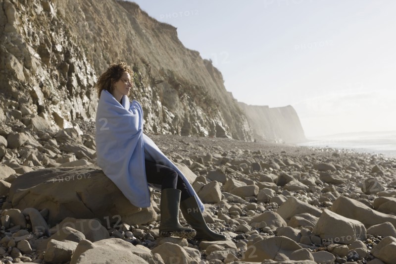 France, Pas-de-Calais, Escalles, Woman wrapped in blanket sitting on rocky beach. 
Photo: Jan Scherders