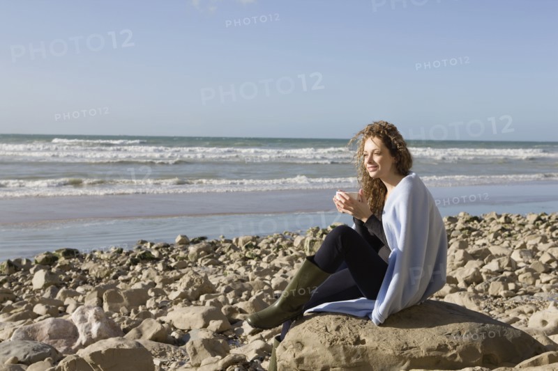 France, Pas-de-Calais, Escalles, Woman wrapped in blanket sitting on rocky beach. 
Photo: Jan Scherders