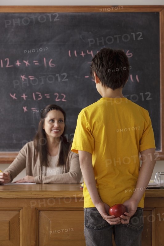 Schoolboy (12-13) holding apple behind back with teacher in background. 
Photo : Rob Lewine