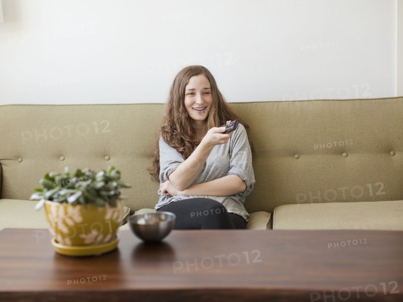 Cheerful young woman sitting on sofa with remote control. 
Photo : Jessica Peterson