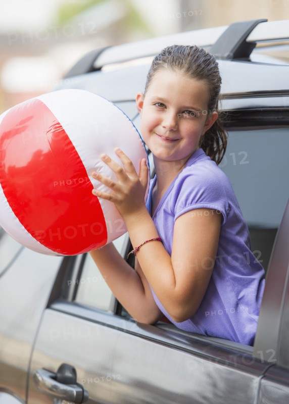 Young girl (8-9) with beach ball peeking through car window . Photo: Daniel Grill