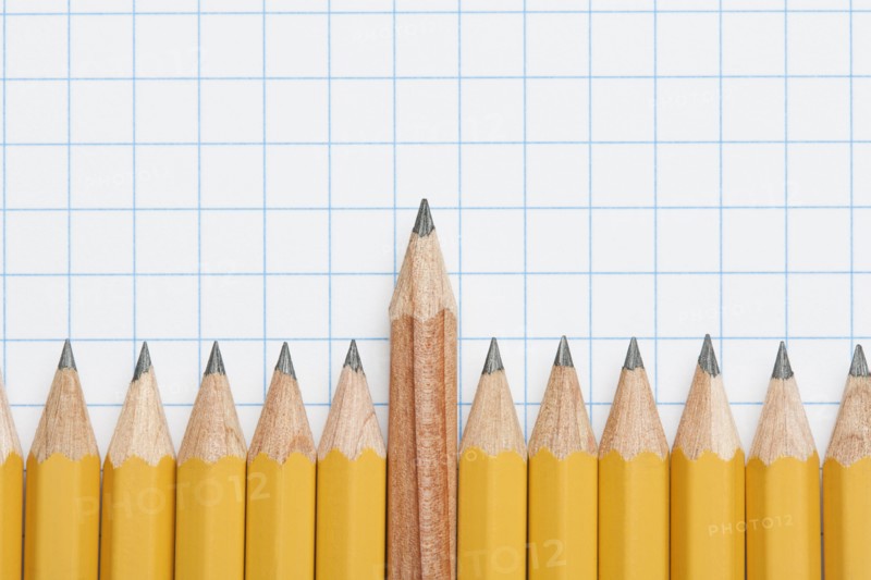 Single wooden pencil rising above row of sharpened pencils on graph paper, studio shot. Photo : Kristin Duvall