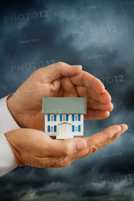 Hands holding model house in front of stormy sky.
