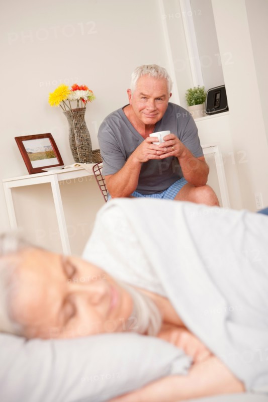 Senior man with coffee cup looking at sleeping wife. Photo: Rob Lewine